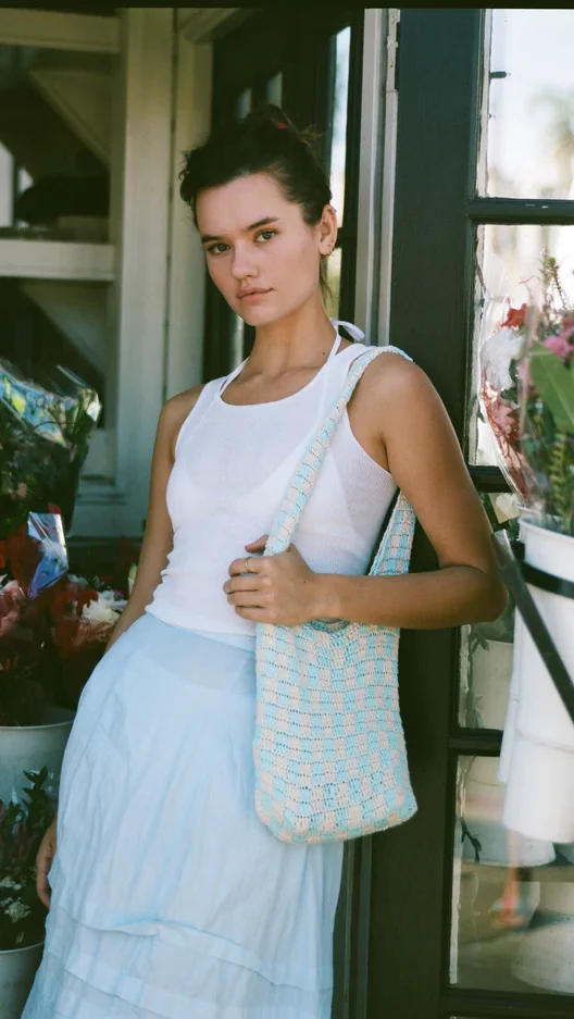 Brunette model holding a blue and white checkered knit tote bag on her shoulder. She is wearing a white tank top, white bikini top, and light blue long skirt.